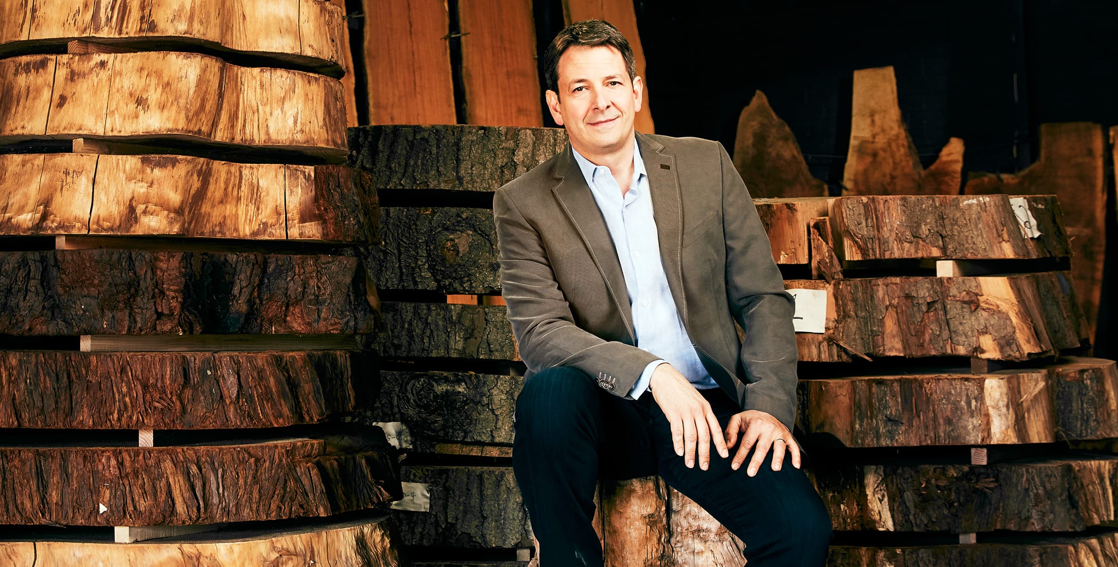 Man in a suit sitting on stacked wooden slabs in a workshop or lumberyard setting.