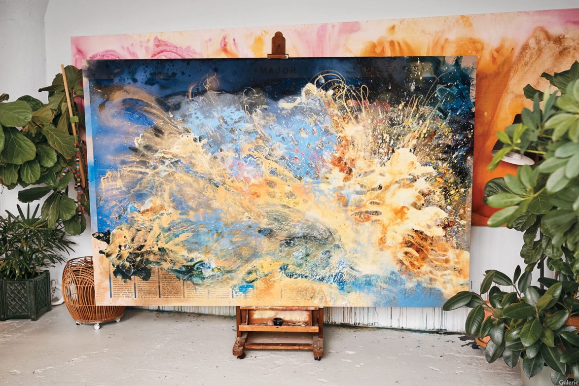 Abstract painting with vibrant splashes of blue, yellow, and orange on a canvas surrounded by potted plants.