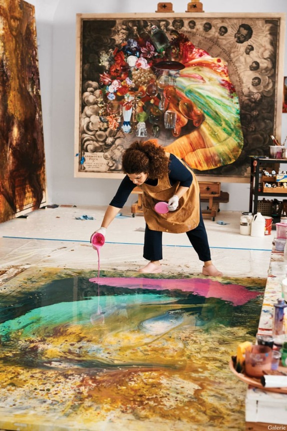 Artist pouring paint on canvas in a studio with large colorful artworks in the background.
