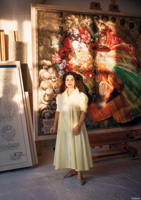 Person in a yellow dress standing in front of a colorful painting in an art studio with architectural drawings nearby.
