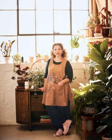 Woman in an artist's apron stands in a brightly lit studio surrounded by plants and natural light.