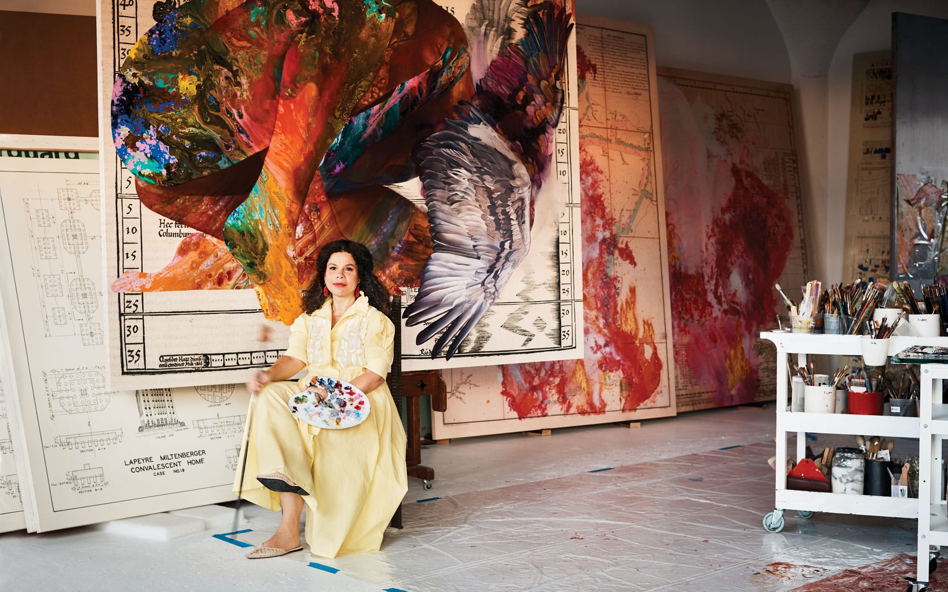 Artist seated in studio surrounded by large colorful abstract paintings and art supplies, holding a palette in her lap.