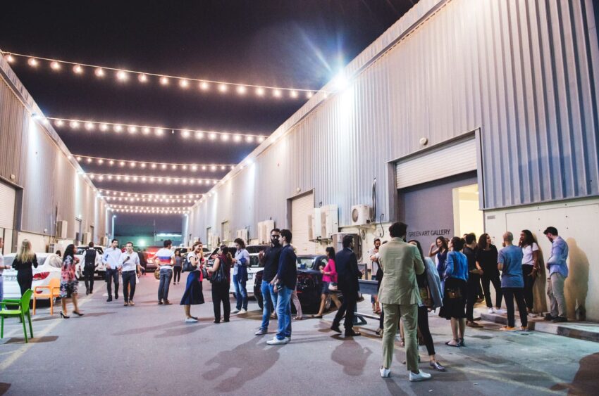 People socializing in a well-lit outdoor gallery with string lights and industrial building background at night.