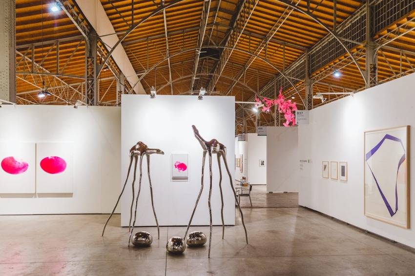 Contemporary art gallery interior with unique sculptures, pink abstract paintings, and framed artwork on white walls.