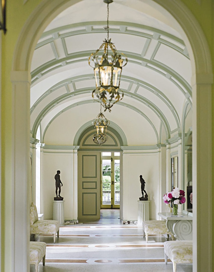 Elegant hallway with arched ceiling, chandeliers, statues on pedestals, and a view of outdoor greenery through large doors.