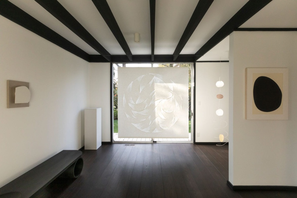 Minimalist room with abstract art, dark wood floor, black beams, and large window with decorative hanging.