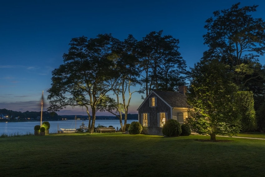 Small house by a lake with illuminated windows and surrounding trees at dusk, creating a peaceful and serene atmosphere.