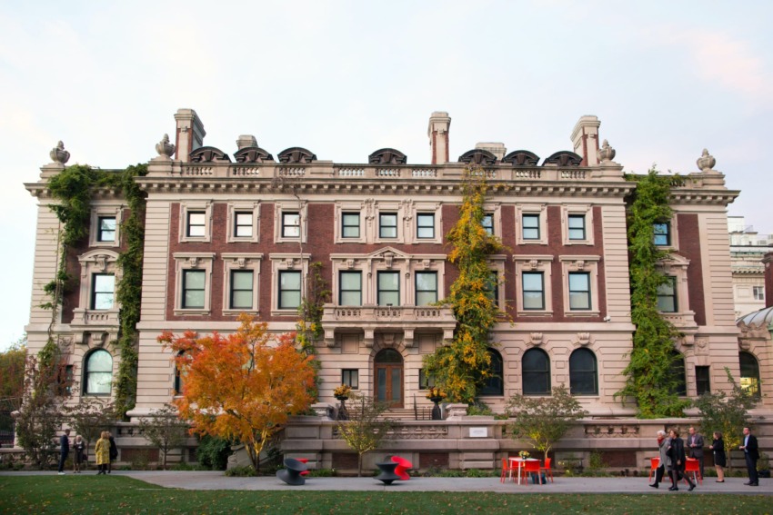 Historic brick building with ivy-covered facade, autumn trees, and people walking in front on a grassy lawn.