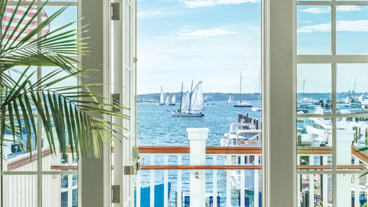 View of a marina through large windows with sailing boats on a sunny day.