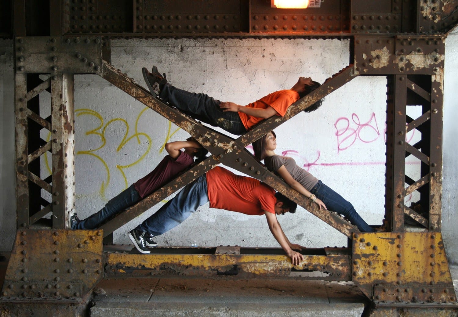 Three people in casual clothing creatively balanced on beams under a bridge, urban art graffiti in the background.