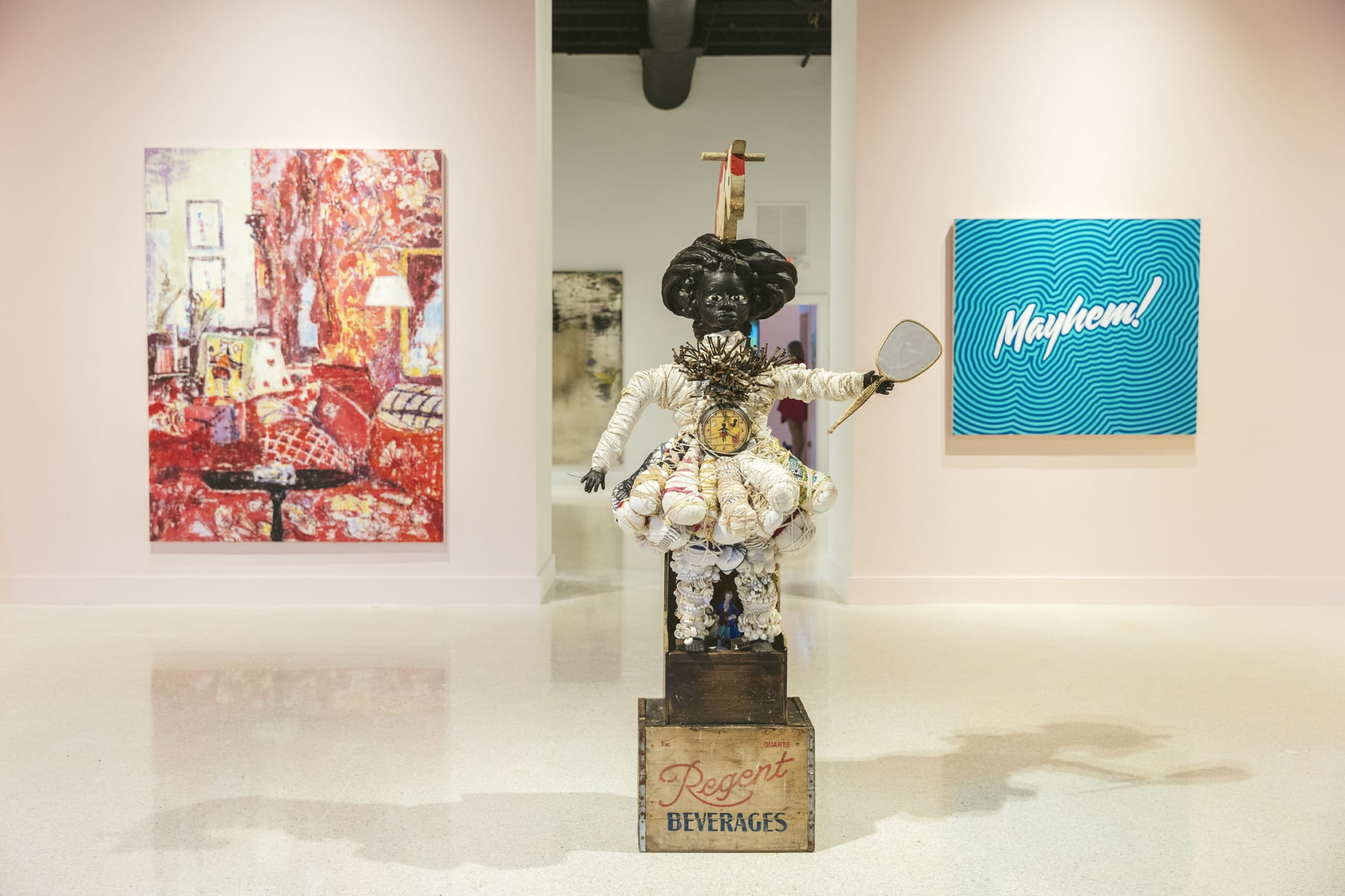 Sculpture with clock and fan on pedestal, surrounded by colorful paintings in an art gallery setting.