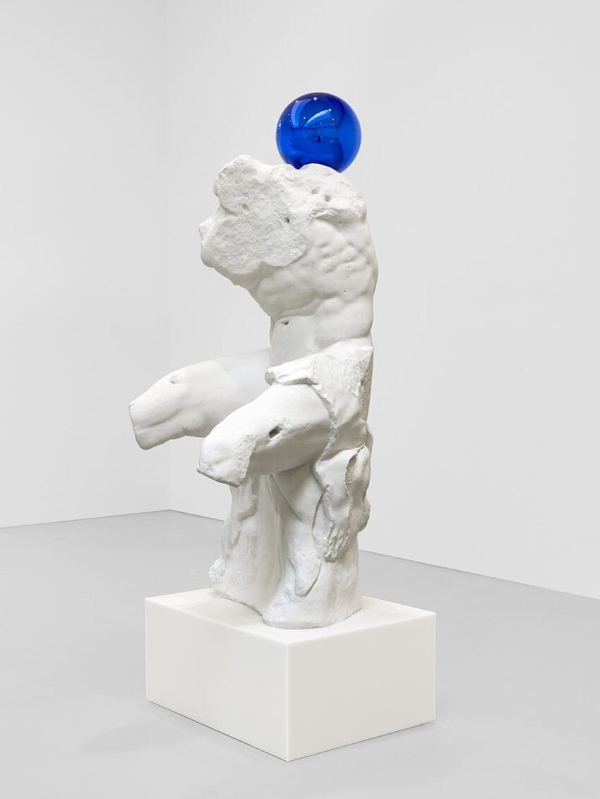 White abstract sculpture with a blue glass sphere on top displayed on a square pedestal in a minimalistic gallery setting.