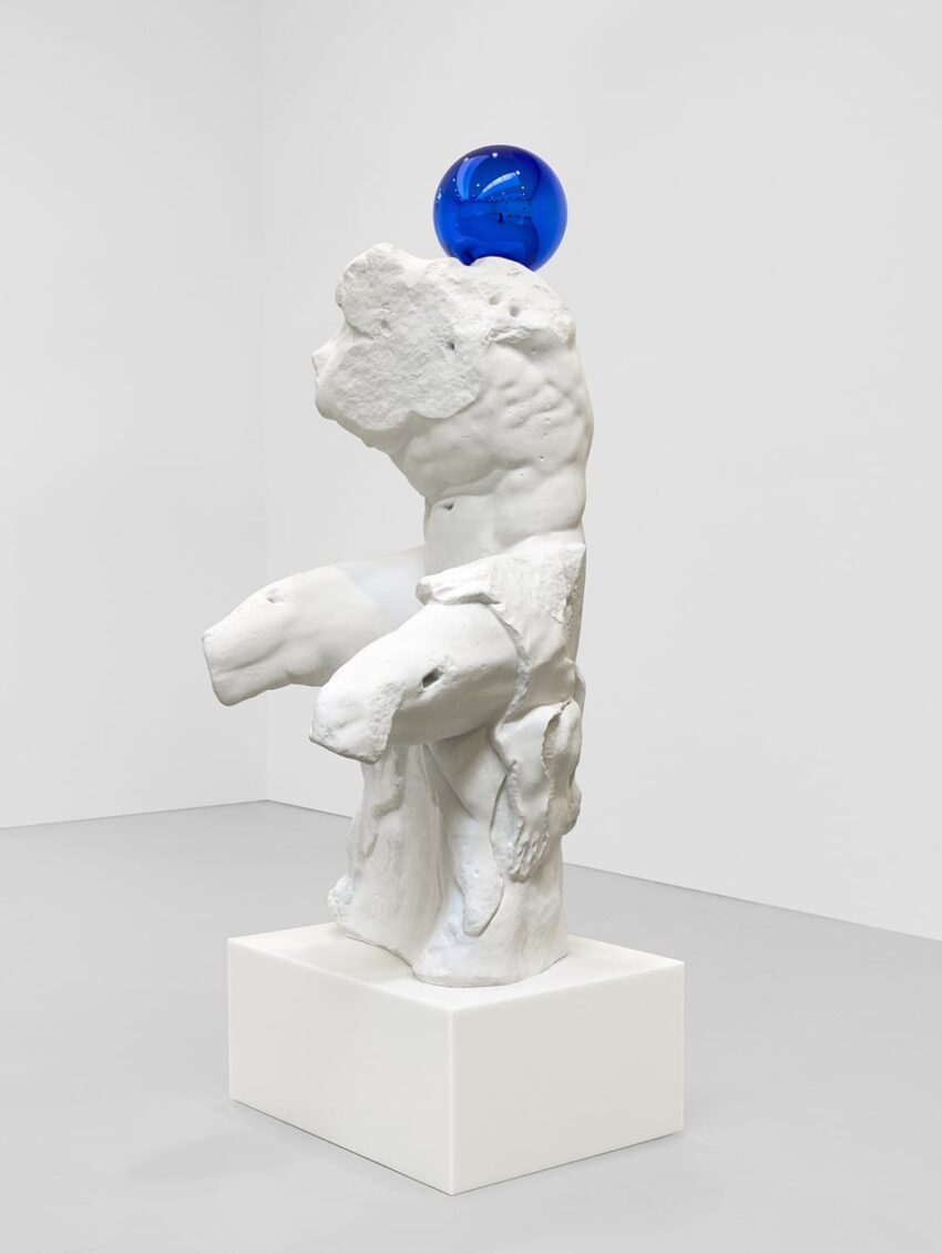 White abstract sculpture with a blue glass sphere on top displayed on a square pedestal in a minimalistic gallery setting.