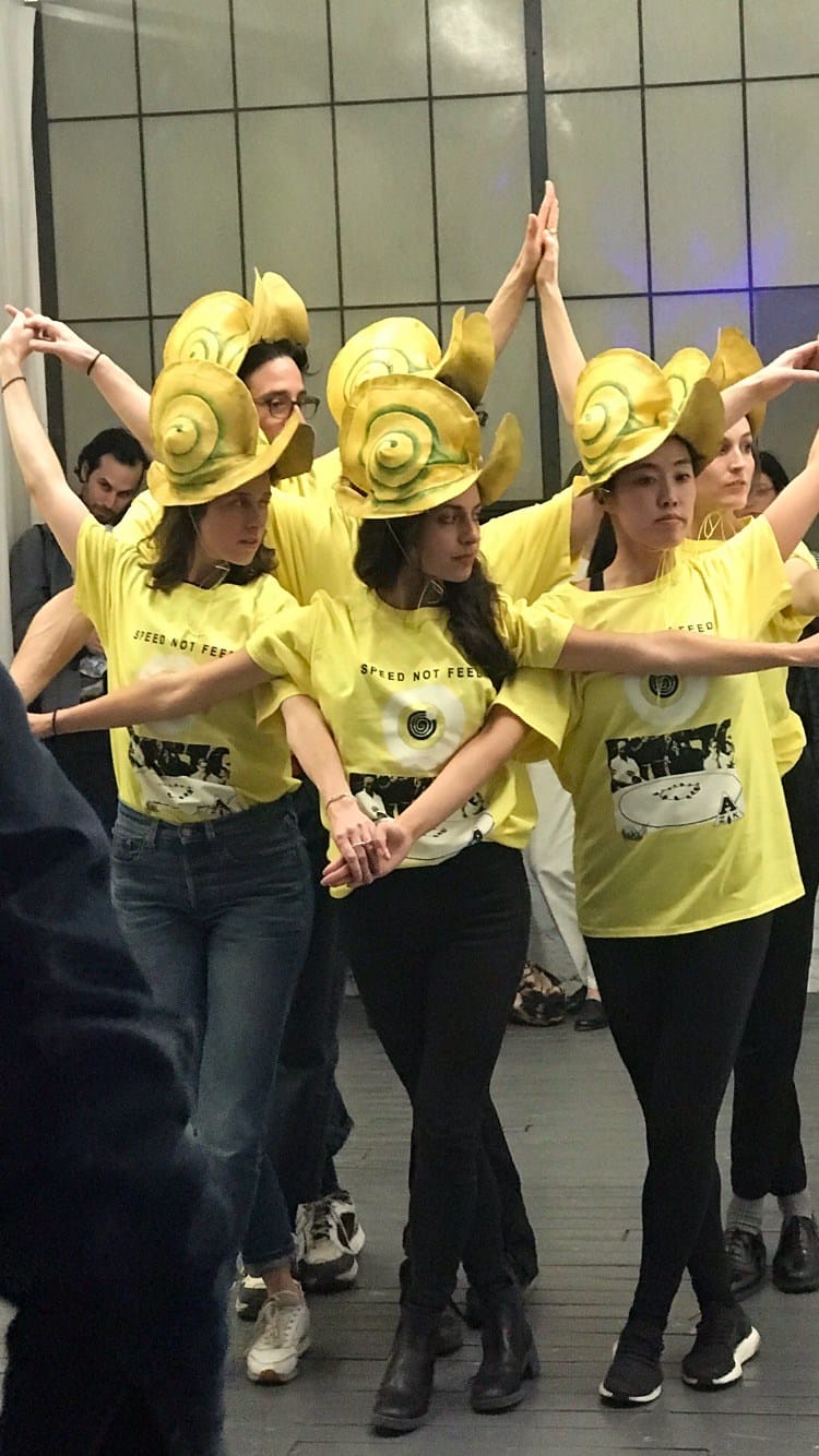 Group of people wearing snail shell hats and yellow shirts dancing in a formation indoors