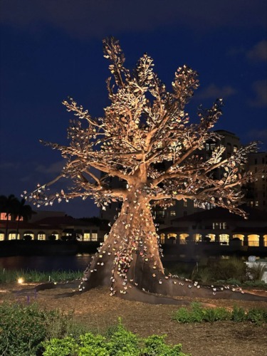 Illuminated sculpture of a tree with intricate metal leaves, set against a night sky, surrounded by building silhouettes.