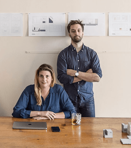 Two people in a modern office setting with architectural drawings on the wall, one seated at a desk and the other standing.