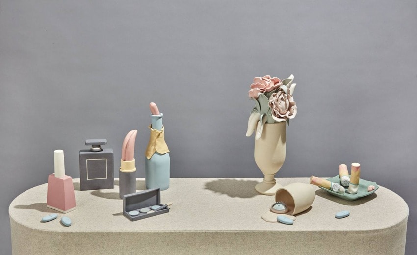 Pastel-colored cosmetic-themed sculptures on a table with a vase of flowers against a plain background.