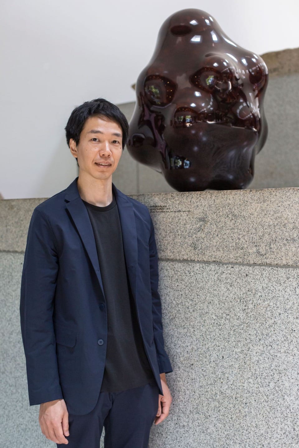 Man in dark suit standing beside abstract glossy black sculpture on granite surface.