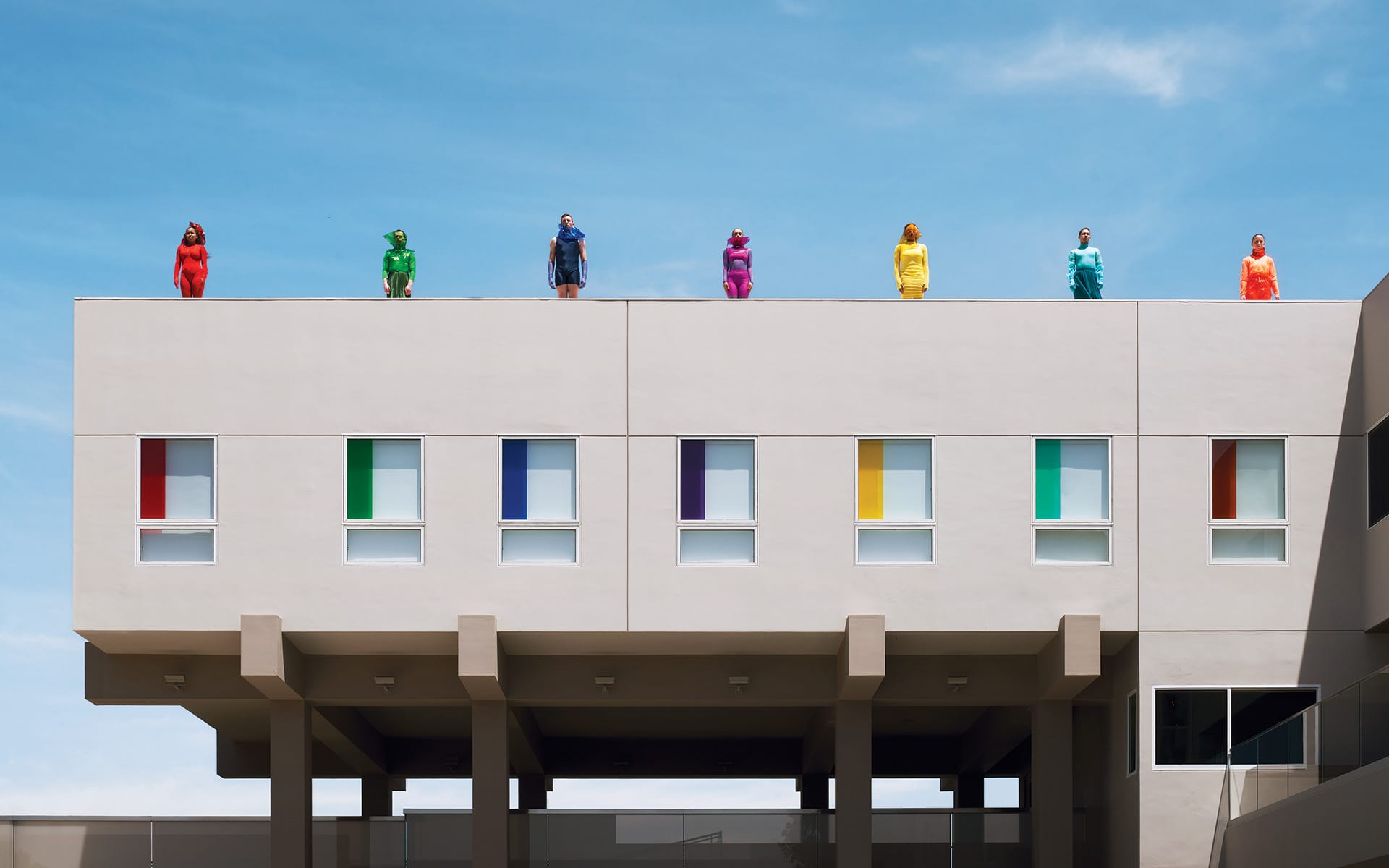 People in colorful outfits standing on a modern building roof with matching rainbow-colored windows under a clear blue sky.
