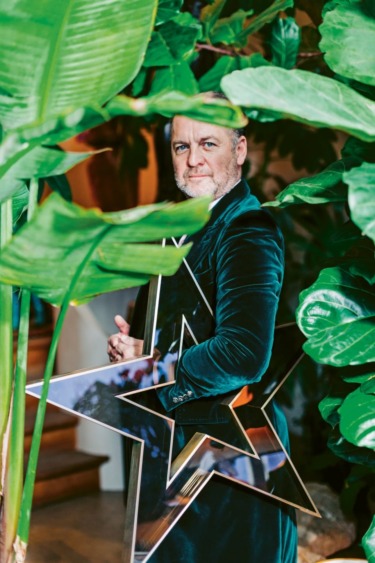 Man in a dark velvet suit holds a large mirror star, surrounded by lush green leaves.
