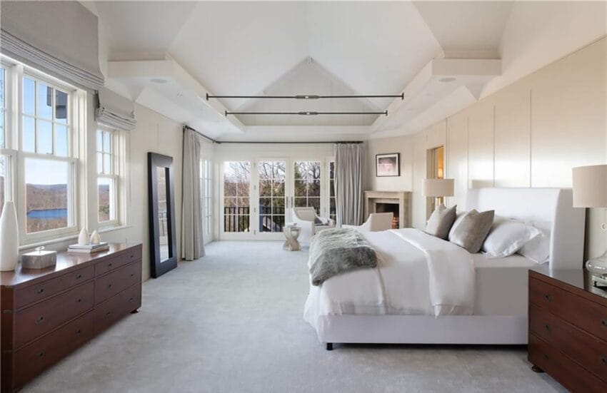 Spacious modern bedroom with large windows, white bed, wooden furniture, and elegant decor.