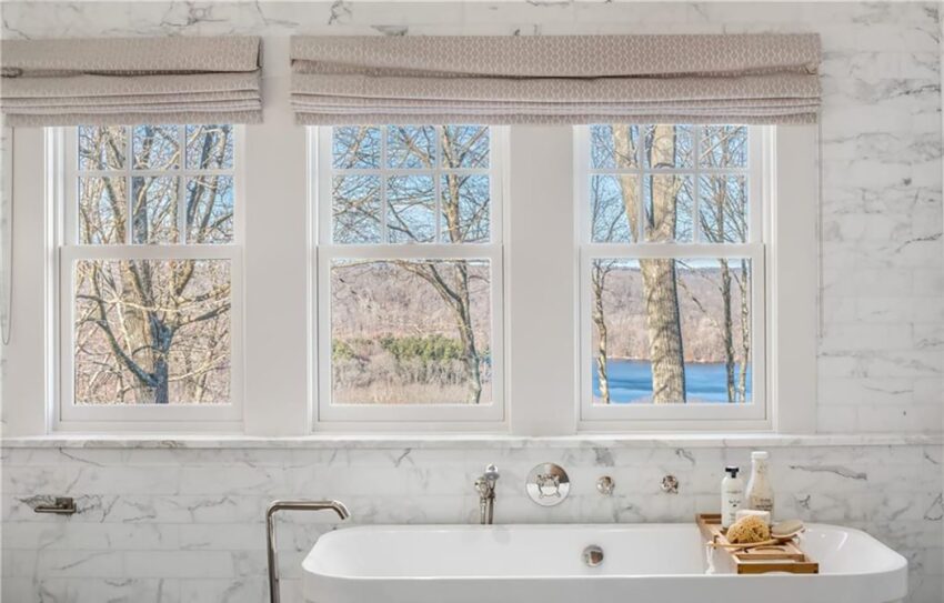 Scenic bathroom with large windows, overlooking trees and river, featuring marble walls and a freestanding bathtub.