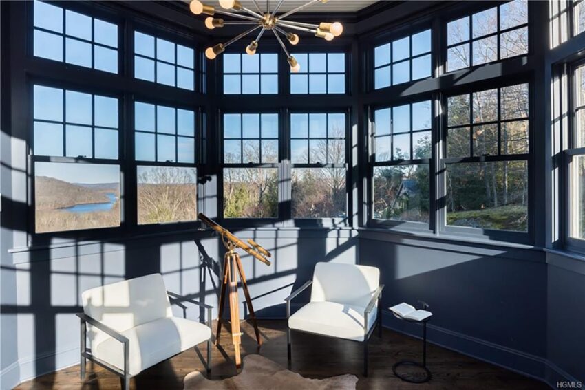 Sunlit room with large windows, telescope on tripod, two white chairs, wooden floor, view of trees and river.