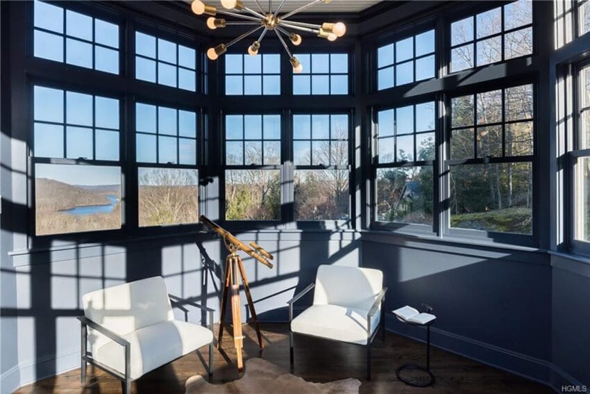 Sunlit room with large windows, telescope on tripod, two white chairs, wooden floor, view of trees and river.