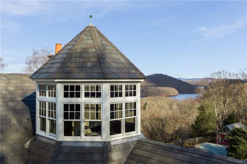 Tudor-style house with slate roof and octagonal tower overlooking a scenic lake and wooded landscape in the background.