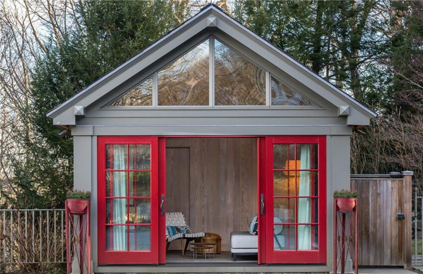 Small modern garden shed with red sliding doors and large windows surrounded by trees.