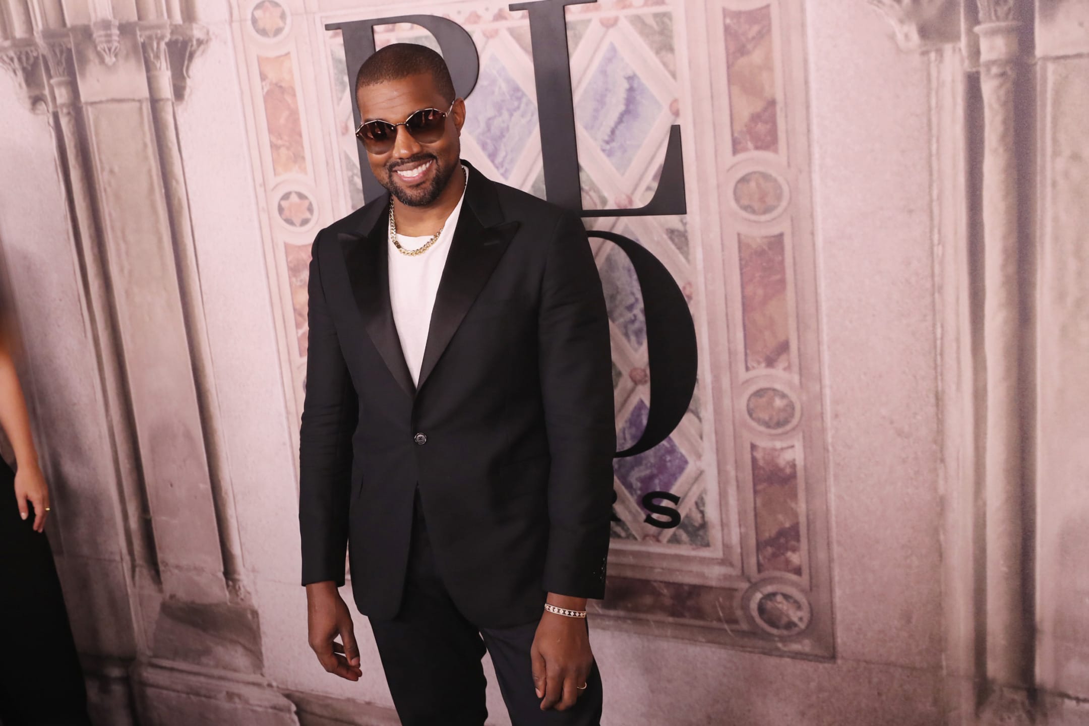 Smiling man in black suit and sunglasses at a fashion event with ornate background.