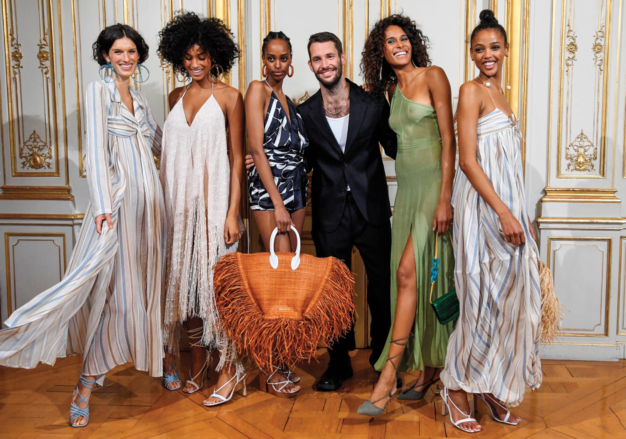 A group of fashion models and a designer in stylish outfits posing together in an elegant room with wooden floor.