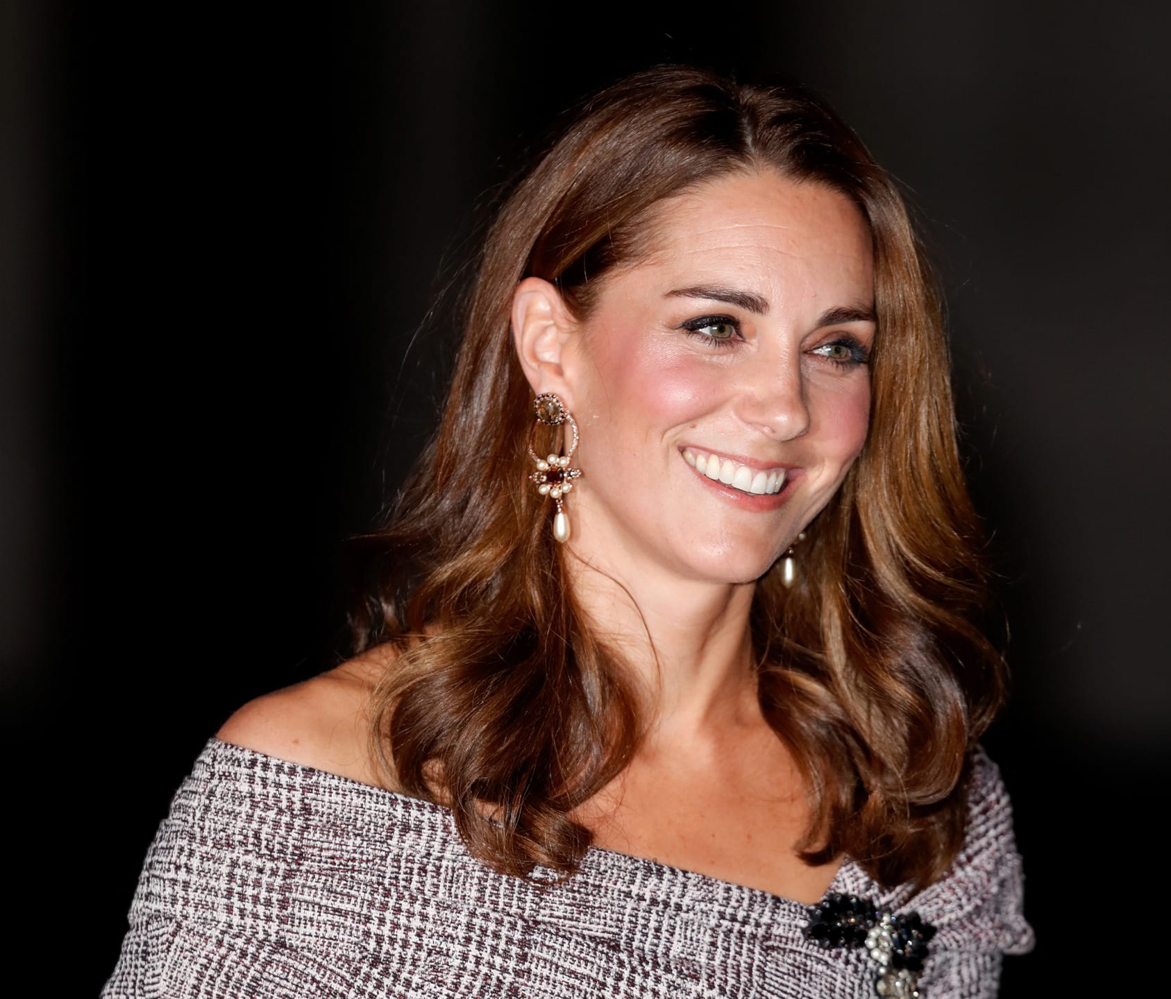 Smiling woman with long brown hair wearing an off-shoulder dress and large earrings in a well-lit room.