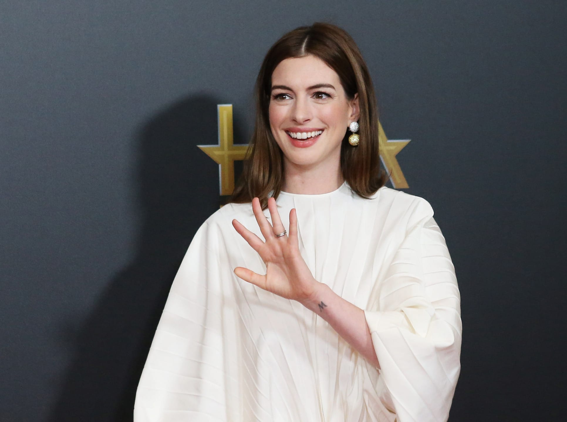 Person in a white outfit waving and smiling against a dark background with gold stars.