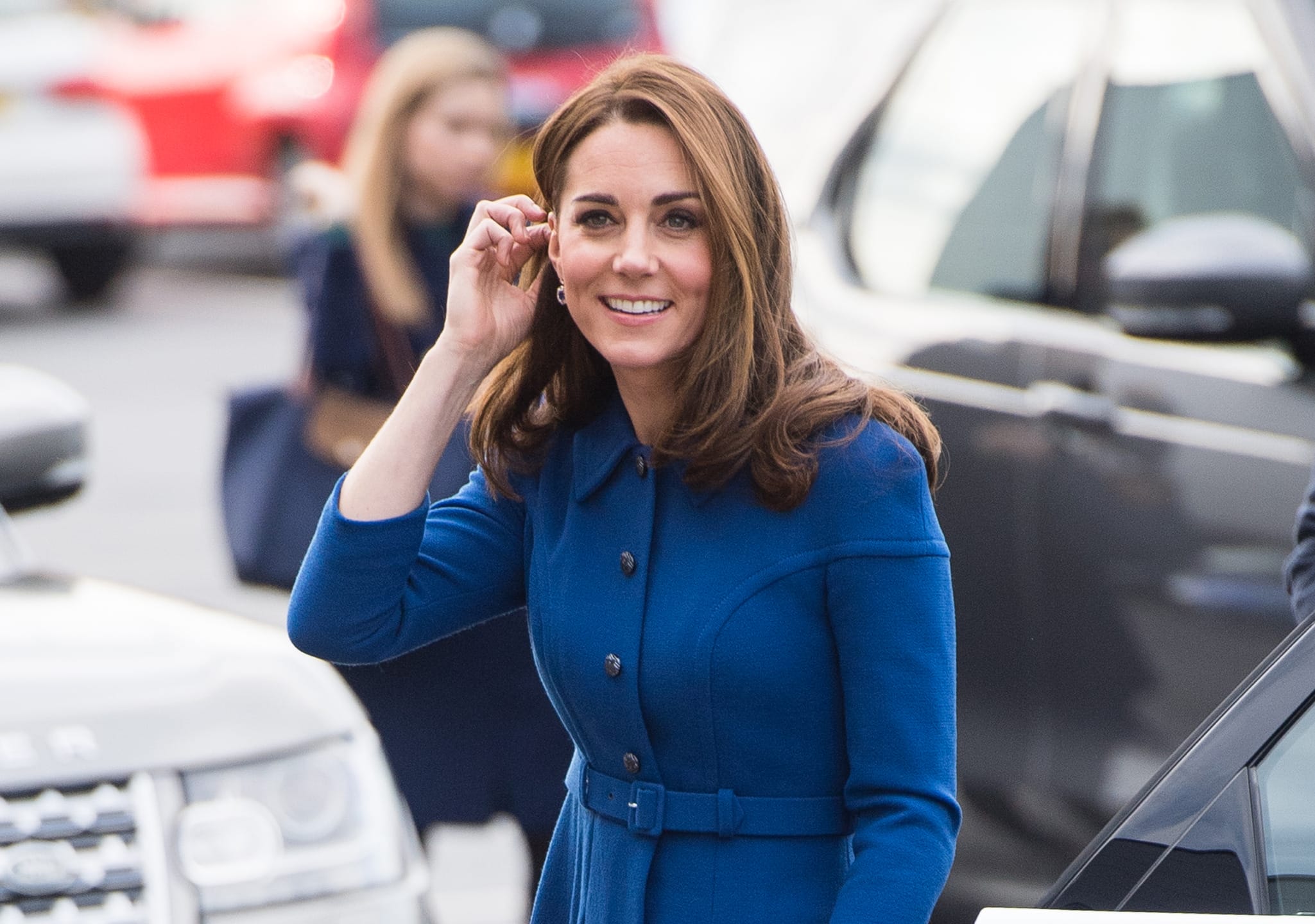 Woman in blue coat smiling while standing outdoors near parked cars.