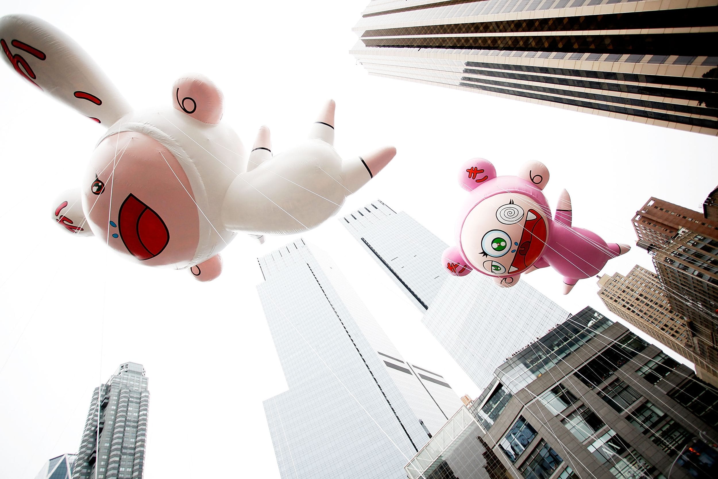 Large character balloons float between skyscrapers at a parade, viewed from a low angle against a cloudy sky.