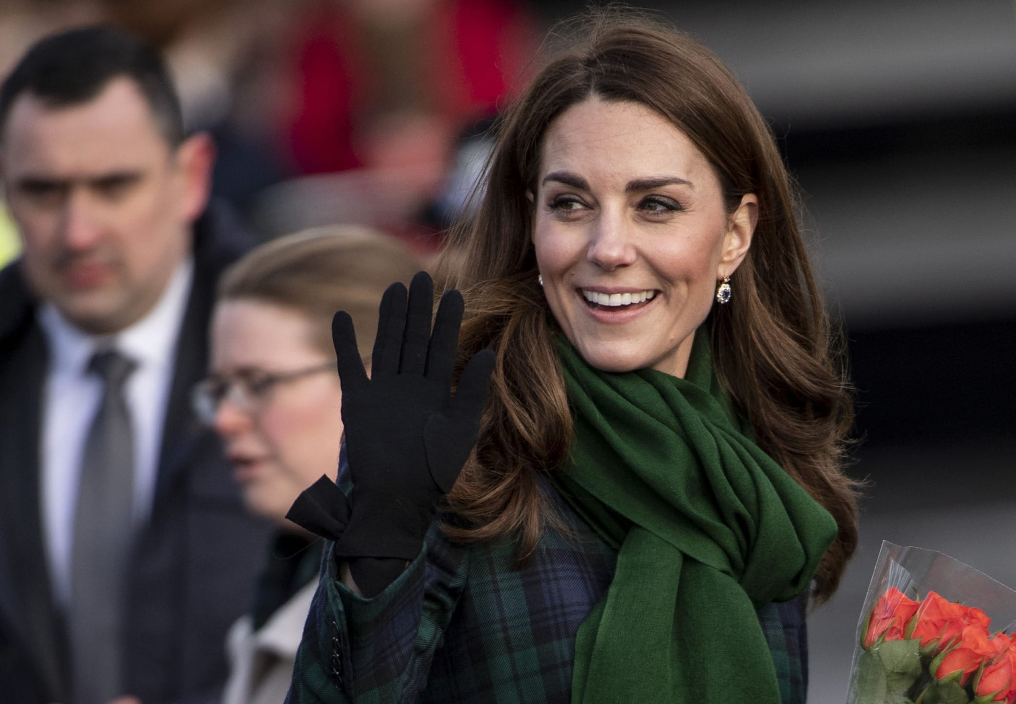 Woman waving while holding flowers, wearing a green scarf and a checkered coat, with people in the background.