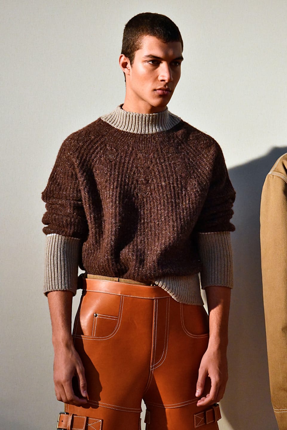 Man in a brown sweater and tan leather pants poses against a light background.