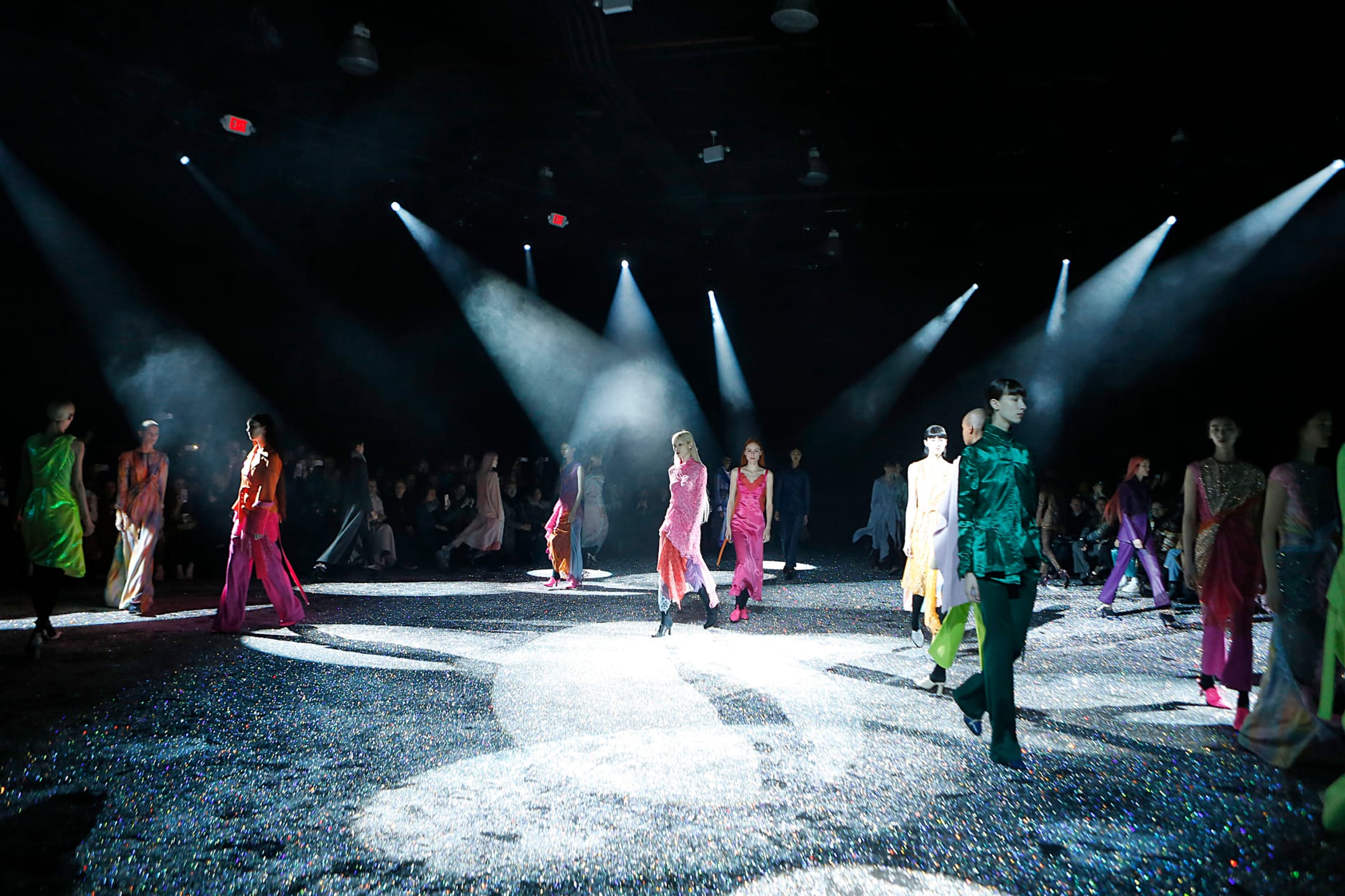 Models walking down a runway in vibrant outfits under spotlight at a fashion show event with an audience watching.