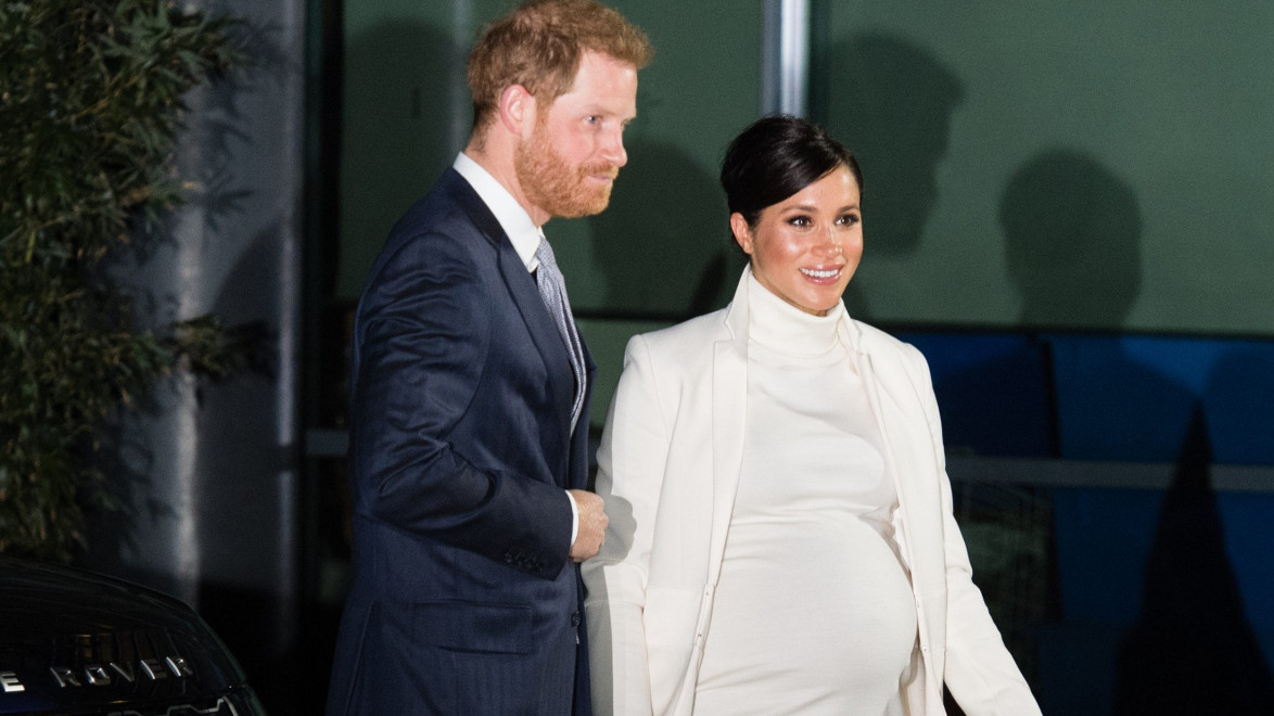 Man in a suit and pregnant woman in a white outfit smiling outside a building.