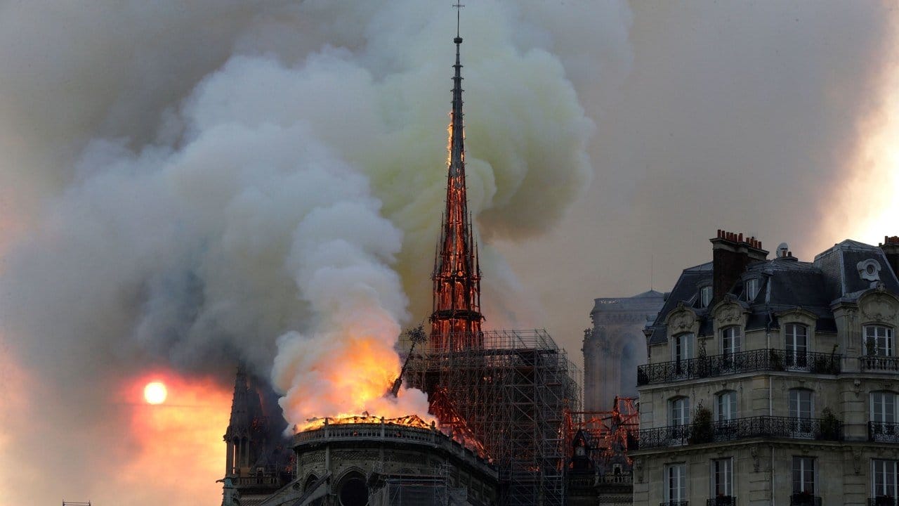 Notre-Dame Cathedral engulfed in flames and smoke during a fire, with the sun visible through the haze in Paris.