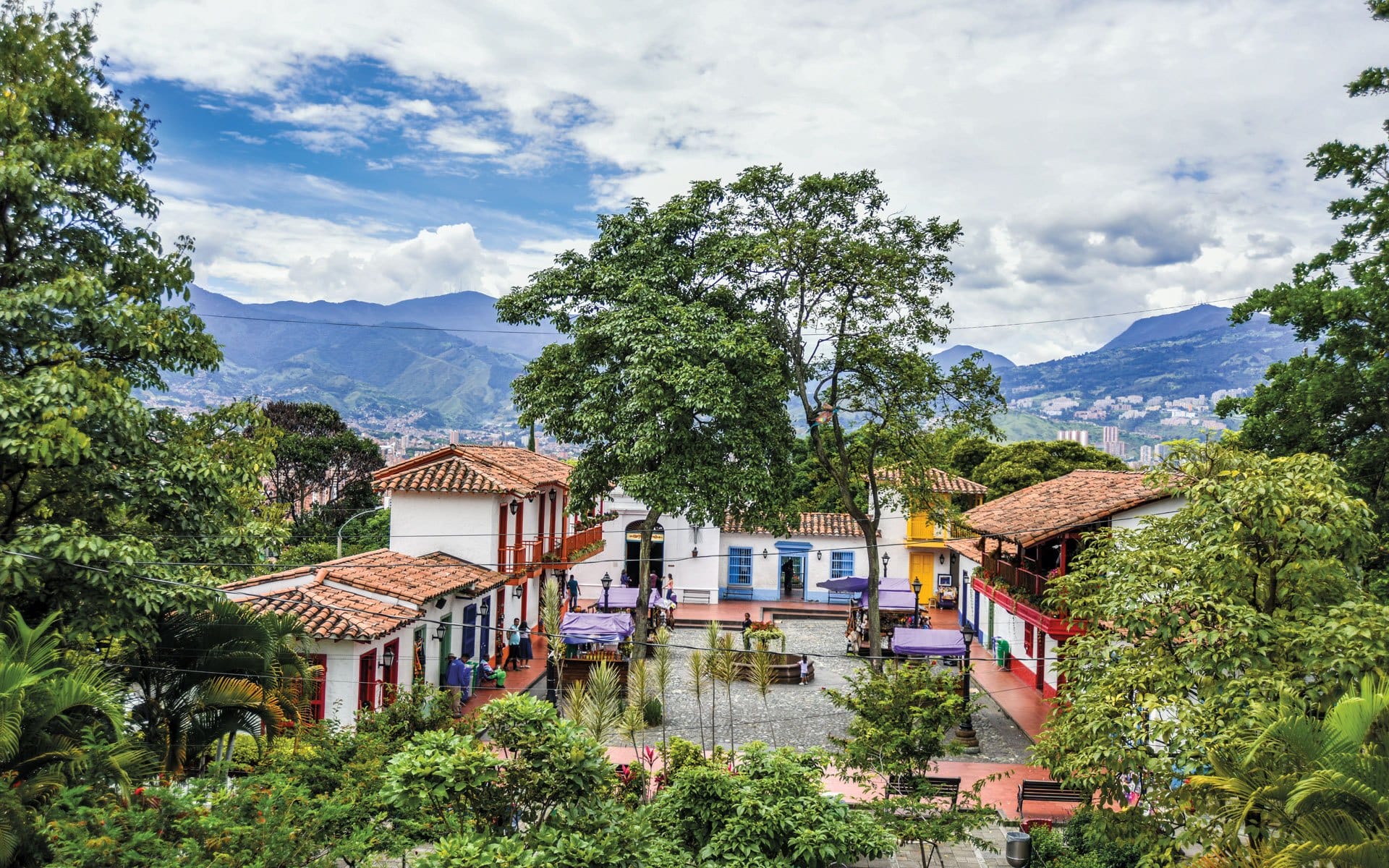 Colorful village square with traditional architecture, lush trees, and mountainous backdrop under a partly cloudy sky.