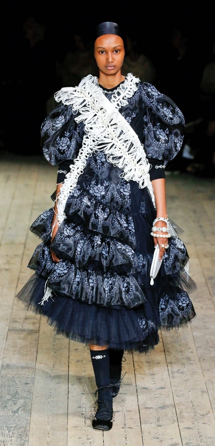 Model in a black textured dress with white accessories walking on a wooden runway, showcasing high fashion design.