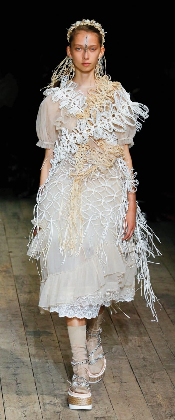 Model wearing an intricate, nature-themed avant-garde dress with layered textures, walking on a wooden runway.