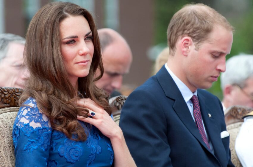 Woman in a blue dress with a sapphire ring sits beside a man in a suit during an outdoor event.