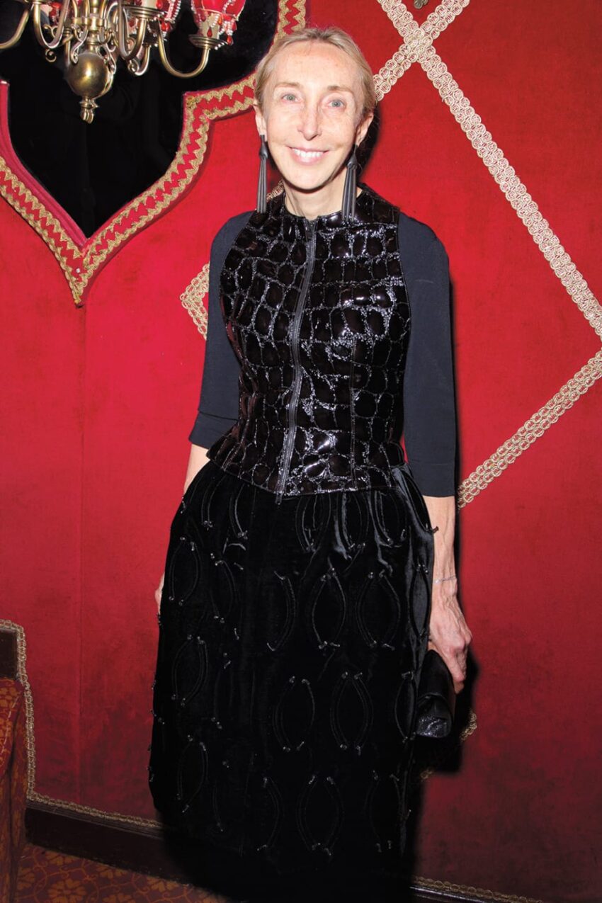 Person in a black velvet dress standing in a red room with ornate decor and a chandelier.
