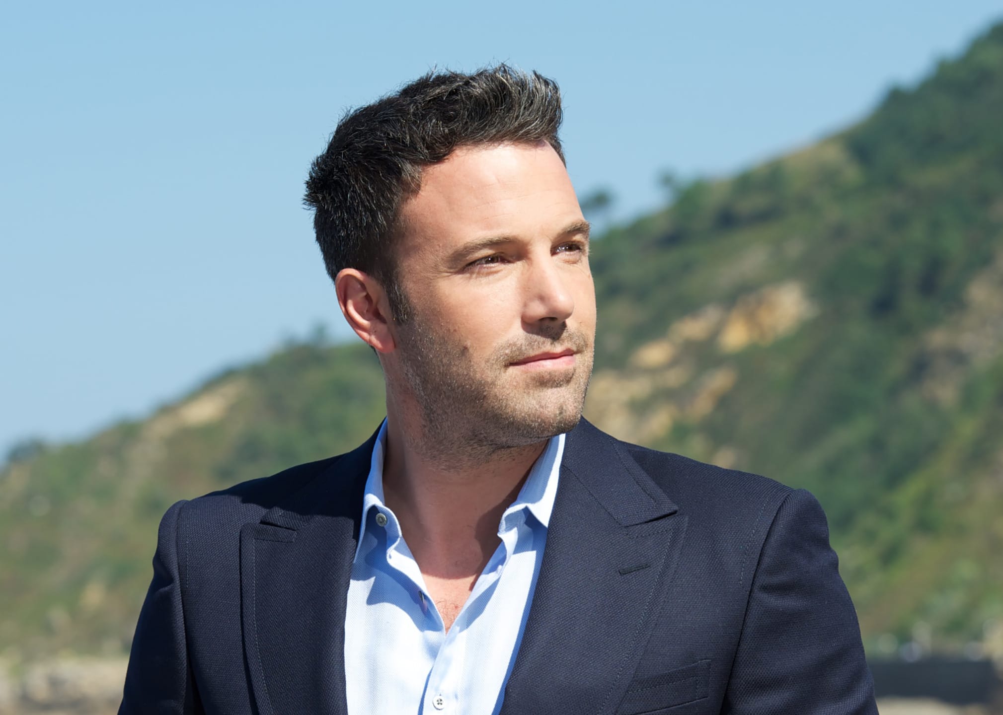 Man in a dark suit with a light blue shirt stands outdoors with a blurred hillside background.