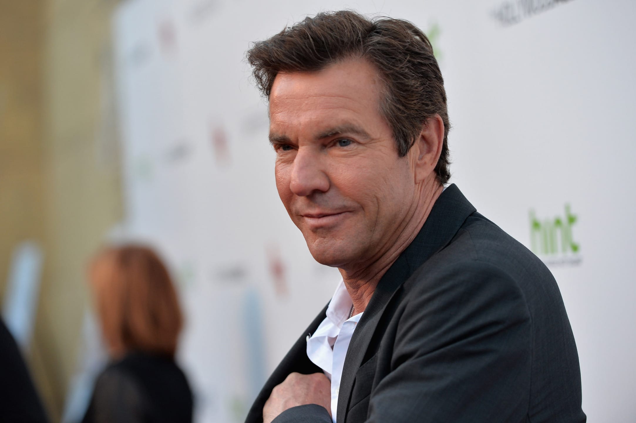 Man in a suit at an event, smiling with a blurred background of people and logos.