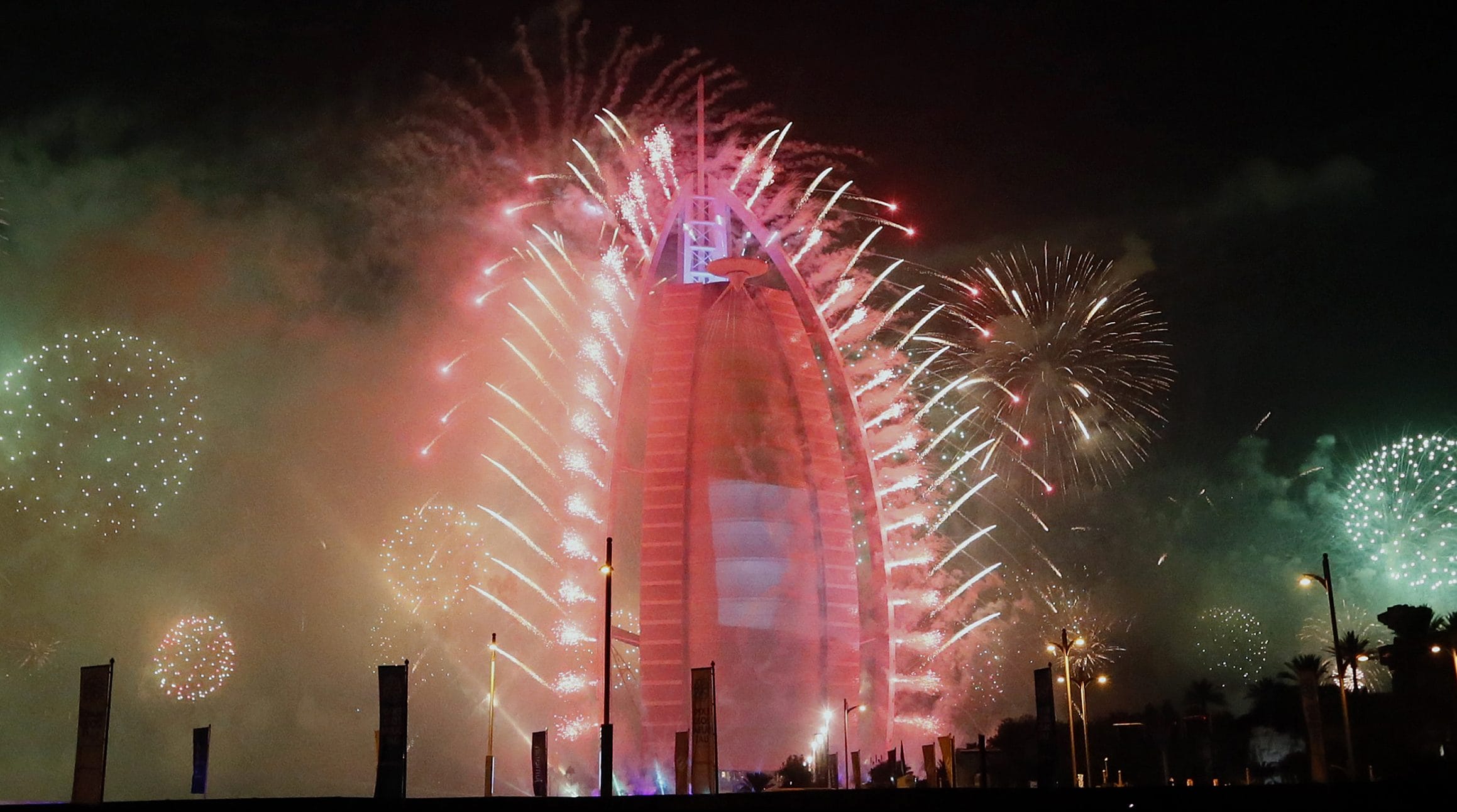 Fireworks display around a brightly illuminated building at night with smoke and colorful lights filling the sky.