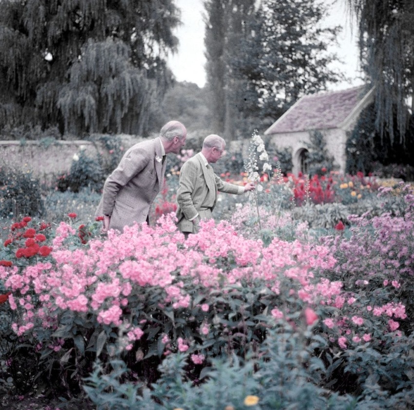 Two men in suits admire flowers in a colorful garden with trees and a small building in the background.